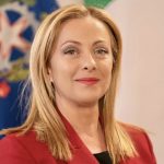 Image of Giorgia Meloni, Italian PM, wearing a red blazer standing in front of flags.